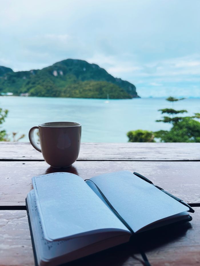 Open journal and coffee cup on a table by a serene beachside view.